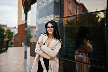 Obraz premium Black hair sexy woman in glasses and coat posed against building with modern windows.