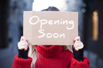 A cardboard tablet close-up with the inscription "Soon opening" in the hands of a young woman in a red sweater.