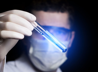 a lab assistant in a mask and glasses holds a test tube with a blue liquid