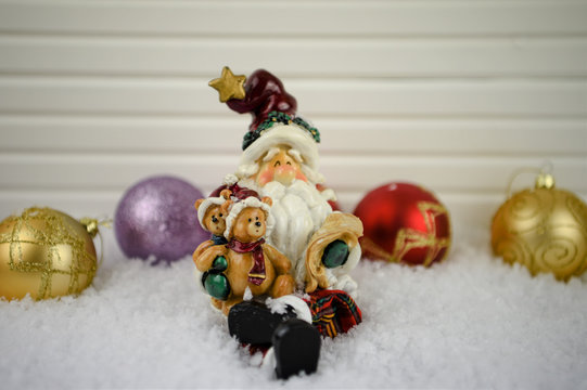 Christmas photography image of traditonal ceramic Santa Claus ornament sitting in snow reading his list with teddy bears and tree bauble decorations in background