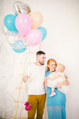 a young family of three people, mom's dad and daughter's one year old stands inside the room. Holding a balloon in her hand,