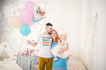 a young family of three people, mom's dad and daughter's one year old stands inside the room. Holding a balloon in her hand,