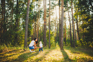 Fototapeta premium beautiful and stylish mother leads the hand of a girl of one year from birth in the park against the background of tall coniferous trees. Family values mom learns to walk child