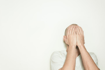 Young depressed man suffering from anxiety and feeling miserable cover his face with his hands and leaning on the white wall