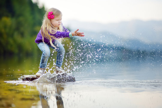 Adorable Girl Playing By Hallstatter See Lake In Austria On Warm Summer Day. Cute Child Having Fun Splashing Water And Throwing Stones Into The Lake.