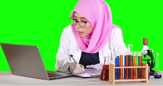 Female scientist writing an experiment report on the clipboard with a laptop and test tube on table. Shot in 4k resolution with green screen background