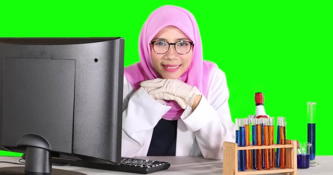 Young female scientist smiling at the camera with computer and test tube on the table. Shot in 4k resolution with green screen background