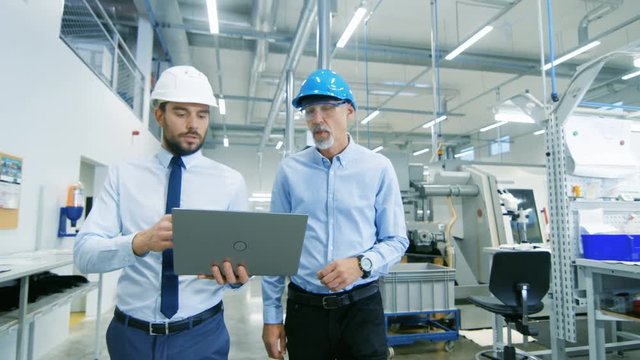 Head of the Project Holds Laptop and  Discusses Product Details with Chief Engineer while They Walk Through Modern Factory. Medium Shot.