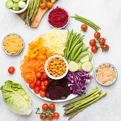 Healthy eating, top view of rainbow buddha bowl, various vegetables, carrot, courgette, cabbage, chickpeas, cucumber and tomatoes, on wooden board on white table, selective focus
