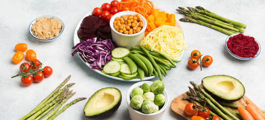 Buddha bowl with ingredients, various vegetables, carrot, courgette, cabbage, chickpeas, cucumber and tomatoes, on wooden board on white table, long photo, selective focus