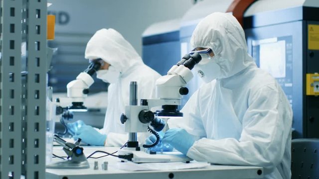 Two Engineers/ Scientists/ Technicians in Sterile Cleanroom Suits Use Microscopes for Component Adjustment and Research. They Work in an Electronic Components Manufacturing Factory. 