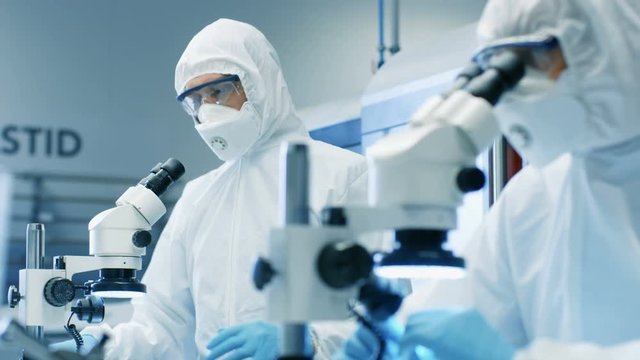 Two Engineers/ Scientists/ Technicians in Sterile Cleanroom Suits  Control Manufacturing Machinery Work and Use Microscopes for Component Adjustment and Research.  