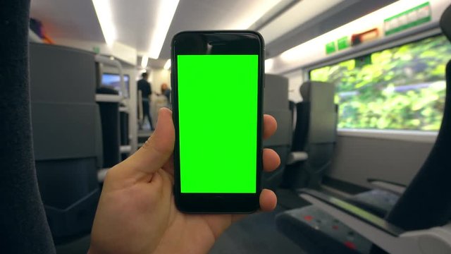 A hand holding (using) a smapthone with a green screen on the train. Close-up shot