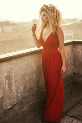 Woman portrait wearing long red, elegant dress on rooftop while looking at cityscape