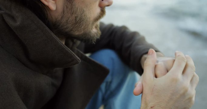 Portrait from the side of the attractive man sitting near the sea and thinking in depressing mood. Cloudy weather. Close up. Outside