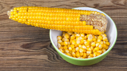 Partially empty corncob and heap of boiled maize. Yellow corn grains in round bowl and partly gnawed cob on a wood background.