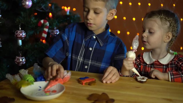 Children cover with multicolored glaze gingerbreads, next to the Christmas tree on the background of electric yellow lights on the eve of the holiday in the evening.