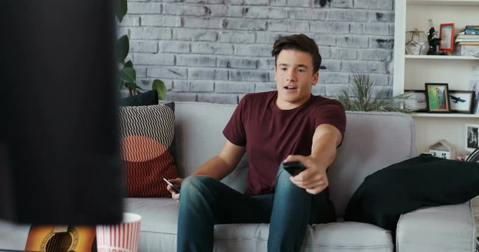 Portrait handsome man turns on the TV set on sofa and uses phone at home modern design playing remote single time apartment couch laziness looking men couch Shot on RED EPIC camera Slow Motion Shot