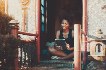 Obraz na płótnie Canvas Charming smiling afro teenage female is sitting on the porch of summer house with tablet pc; young cheerful Brazilian woman with curly hair working with digital pad while sitting on veranda of pagoda
