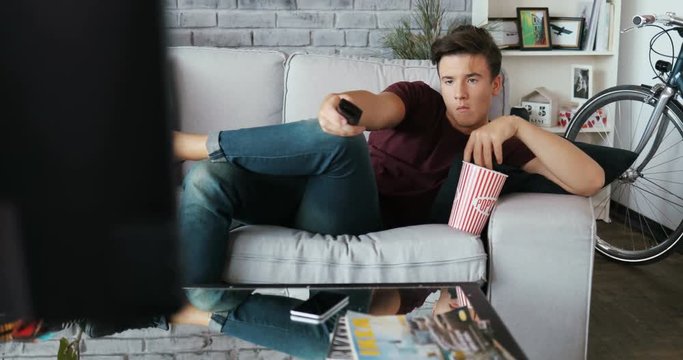 The boring young guy is lying on the couch with remote in his hands to find something watching TV eats popcorn at home modern design looking men Shot on RED EPIC camera Slow Motion Shot