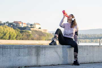 Young girl with headphones on her head sitting and drinking water. She is resting after jogging at the city with river and old fortress at background 