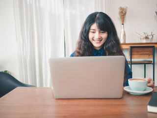 Young asian woman sitting in coffee shop at wooden table, hand holding coffee cup, Using laptop browsing internet, chatting, blogging, shopping onlie.