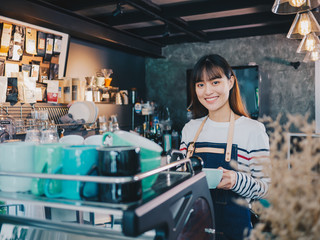 Young asian woman barista with a smile, holding a glass cup of coffee at bar counter.