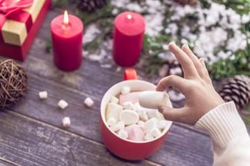 Obraz na płótnie Canvas Christmas still life. Children's hands hold big marshmallows. A cocoa cup from marshmallows on a wooden table against the background of the New Year's still life. Selective focus