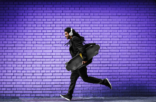 Handsome young dreadlocks skater with headphones running in a suit near the violet wall on the street.