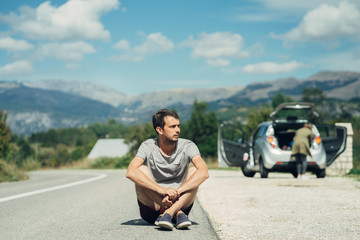 Fototapeta na wymiar Young man is sitting in front of car on empty road. Road trip stop. Road trip break.