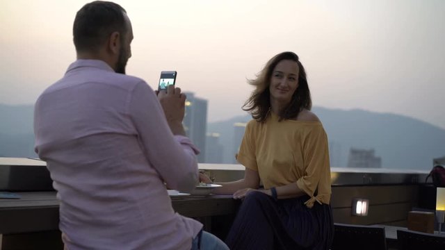 Man taking photo of his beautiful girlfriend sitting on terrace in bar
