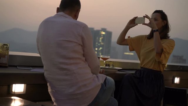 Woman taking photo of her boyfriend with cocktail sitting on terrace in bar
