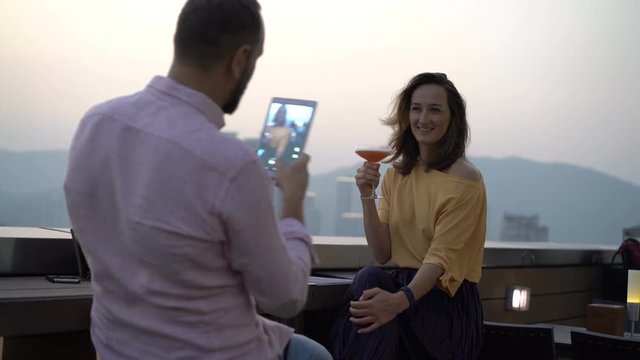 Man taking photo of his beautiful girlfriend with cocktail sitting on terrace in bar
