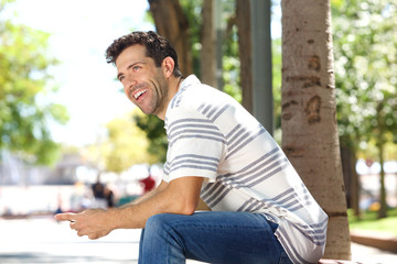 Handsome young man sitting outdoors with mobile phone and laughing