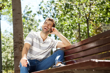 Laughing young man sitting outdoors on bench and using mobile phone