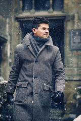 A stylish young man in a warm gray coat walking in the street during the snowfall.