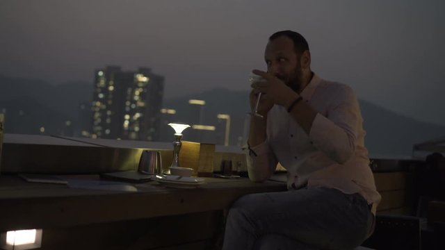 Young man talking on cellphone and drinking cocktail on terrace in bar at night
