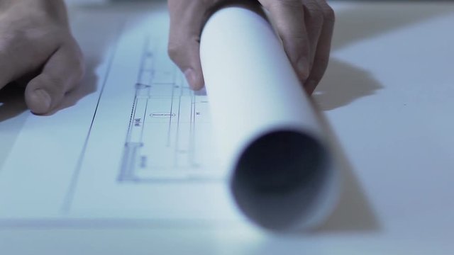 Engineer unrolling drawing of building on table at design office, slow-mo