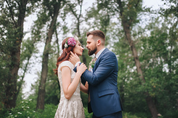 The bride with a wreath on her head gently embraces a bearded bridegroom in a summer forest.