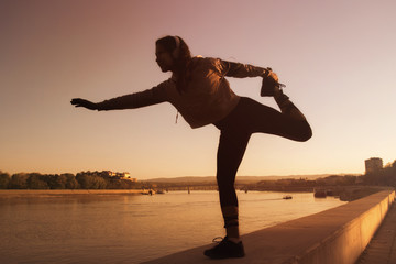 Silhouette of young fitness girl at sunset making pirouette with river and bridge on background © pucko_ns