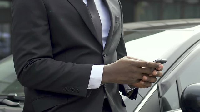 African American man in business suit typing message standing near his car