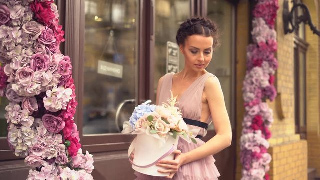 attractive young woman in evening dress standing at the entrance to a flower shop. the girl is holding a cardboard hat box with flowers in cancases. a large bouquet of flowers. the girl demanstrates a