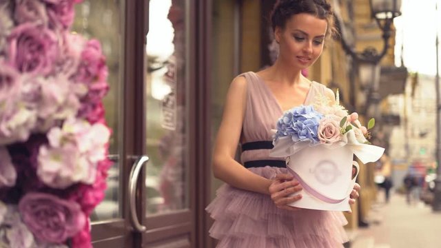 attractive young woman in evening dress standing at the entrance to a flower shop. the girl is holding a cardboard hat box with flowers in cancases. a large bouquet of flowers. the girl demanstrates a