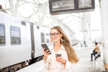 Portrait of a young woman using smart phone at the railway station with information board on the...