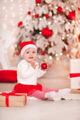 Obraz na płótnie Canvas Happy baby girl 1 year old holding Christmas ball sitting on floor over Christmas background in room. Looking at camera. Holiday season.