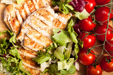 Close up on grilled chicken breast on fresh cut salad next to a bunch of tomatoes on a wooden board