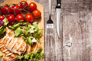 Grilled chicken breast on fresh cut salad next to a bunch of tomatoes on a wooden board. A fork with a knife is next to the board