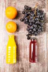 Orange and grape juice in bottles next to two oranges and a grape bunch on wooden background. Healthy fresh lifestyle