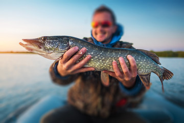 Happy amateur angler holds pike fish with lake on the background. Tilt shift effect applied