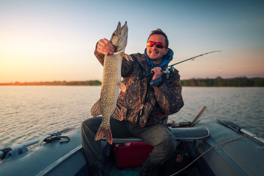 Happy amateur angler holds Pike fish sitting in the boat on the lake during sunrise